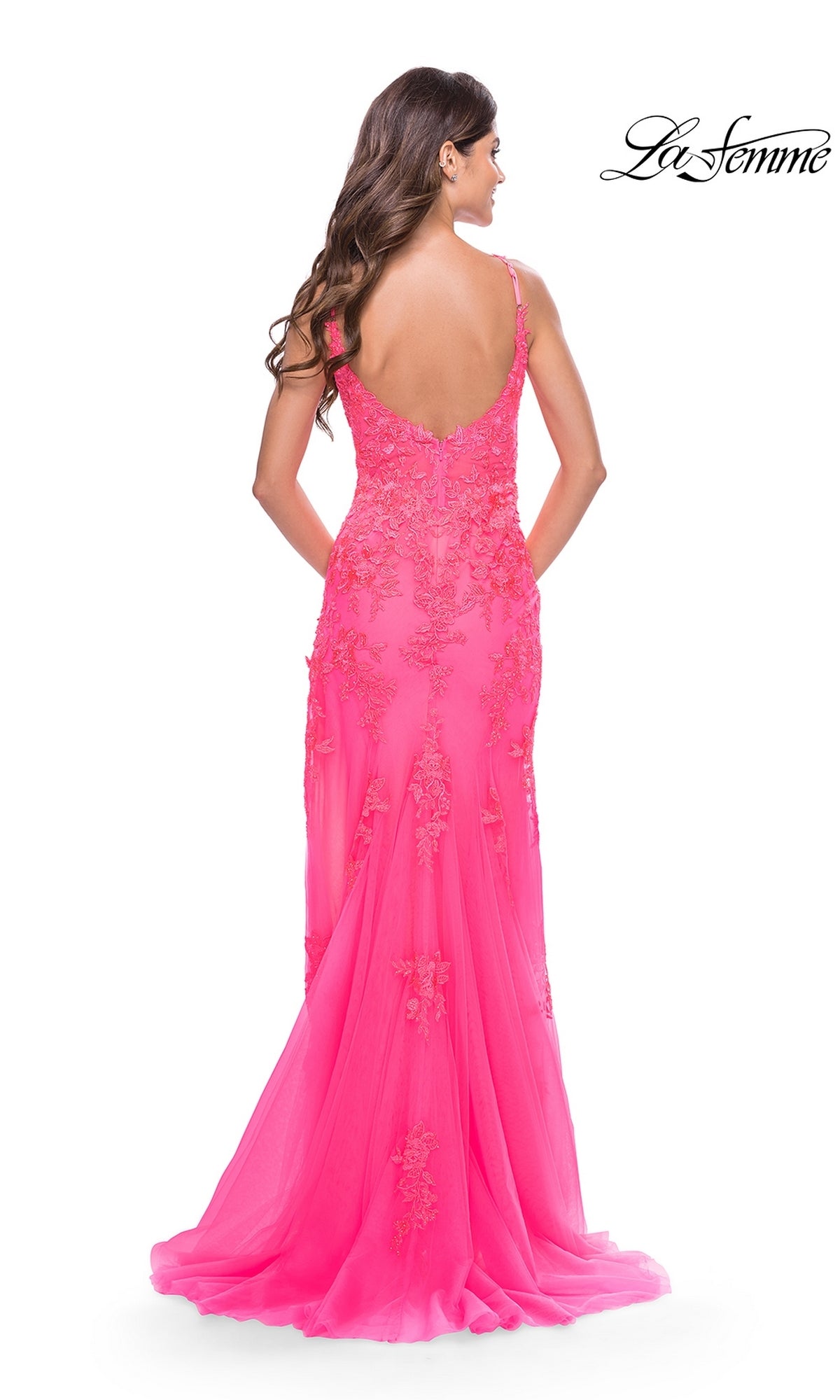 Woman in a sheer-bodice, bright pink, floor-length La Femme Long Lace Prom Dress 31125 with a low back stands facing away from the camera, hands held behind her back.